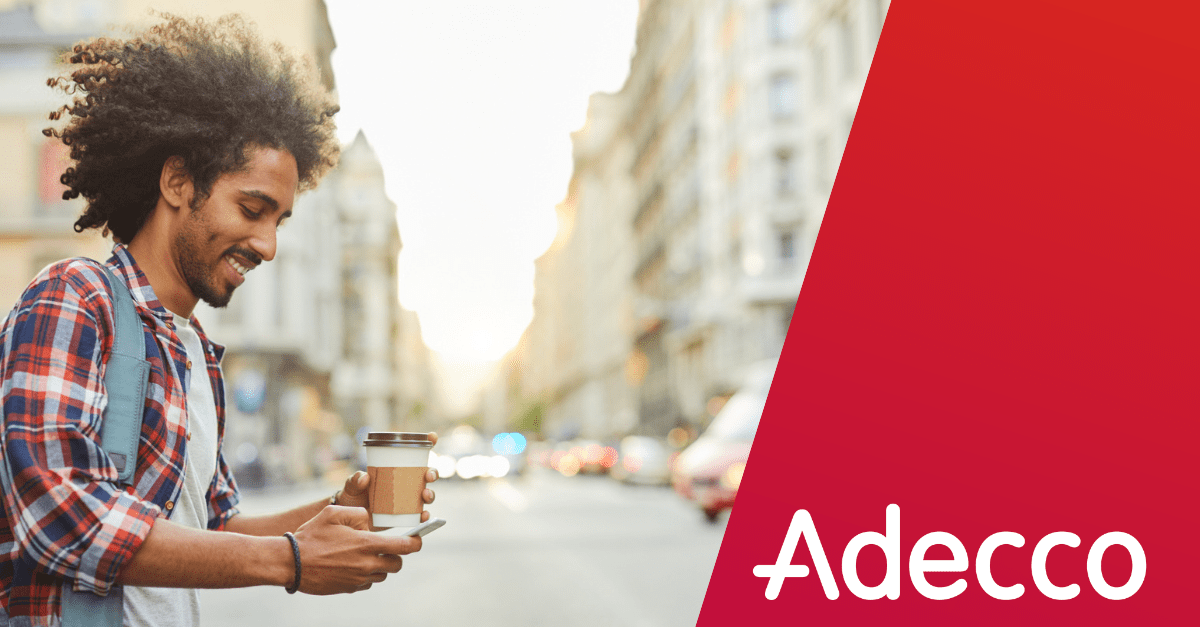 A smiling individual walks down a city street, looking at his phone and holding a cup of coffee